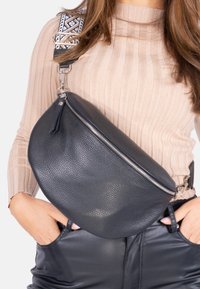 Schwarze Leder-Umhängetasche mit einer strukturierten Oberfläche, silbernem Reißverschluss und einem gemusterten Riemen. Kompakte, abgerundete Form.