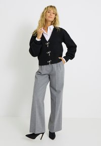 Cardigan noir en peluche, court, avec un col en V et des accents de nœud argentés, associé à un pantalon large gris et des escarpins noirs à bouts pointus.