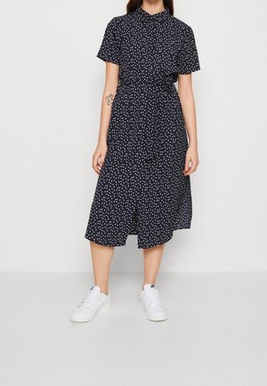 Woman wearing a navy blue, short-sleeve, button-up dress with white polka dots and white sneakers, standing against a plain background.