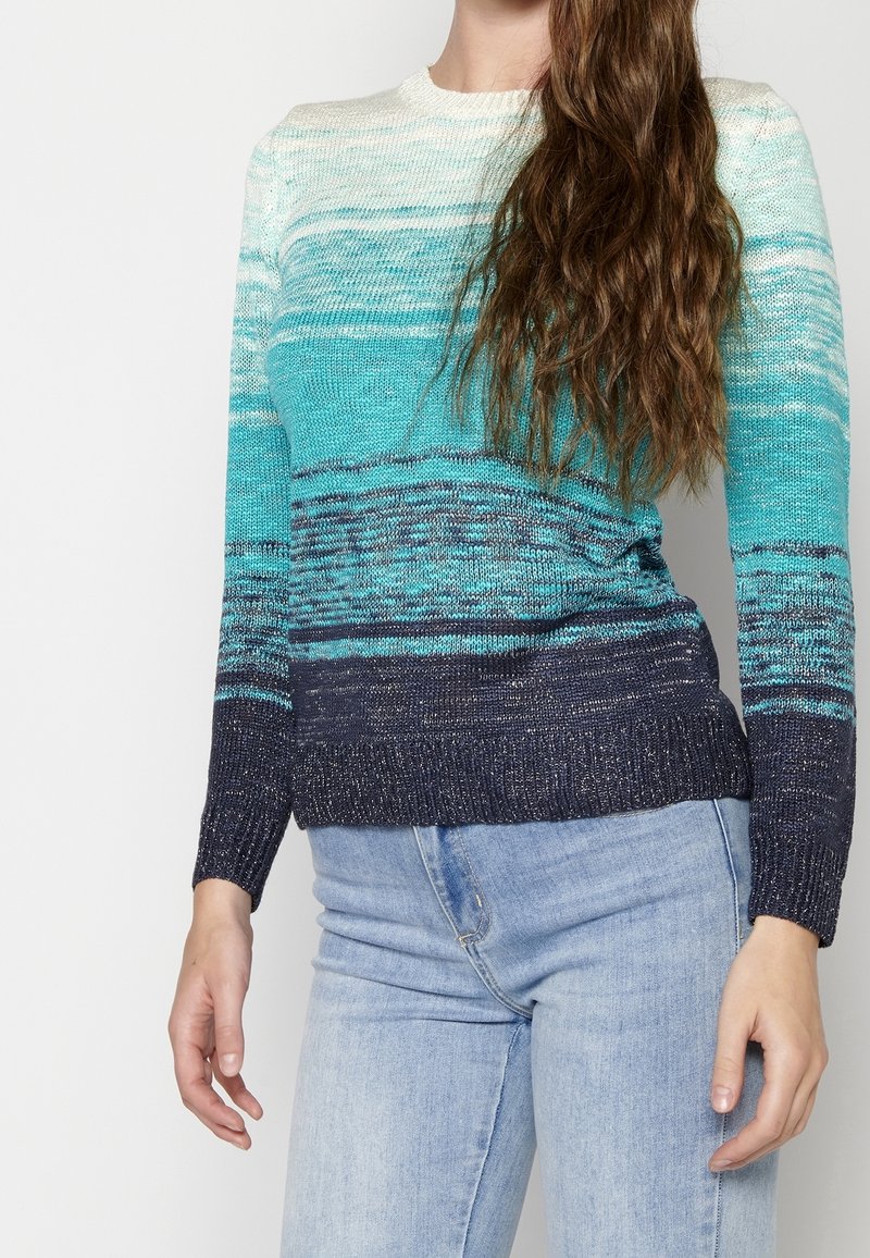 Femme portant un pull en maille à manches longues dans des tons de bleu et sarcelle avec un jean bleu clair, debout devant un fond blanc.