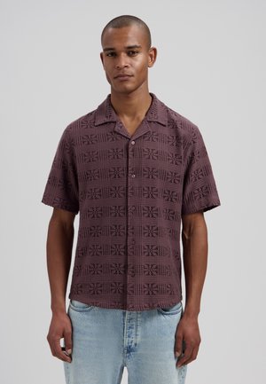 Man draagt een bruin patroon overhemd met korte mouwen en knopen, en lichtblauwe spijkerbroek, staand tegen een effen achtergrond.