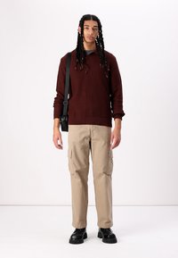 Bordeaux ribbet sweater, beige cargo bukser med sidelommer, sorte chunky støvler, model med fletninger står mod en hvid baggrund.