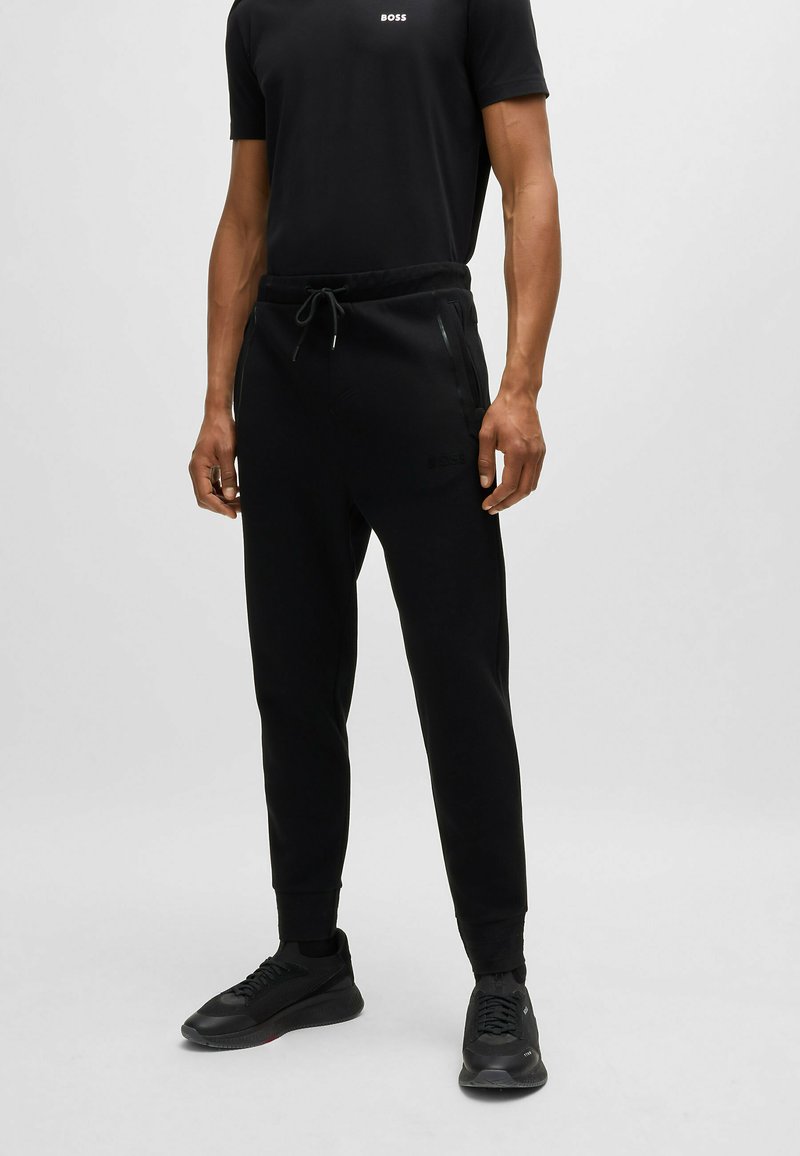 Pantalones de chándal negros con cintura elástica y cordón, que cuentan con bolsillos laterales con cremallera y un logo sutil en el muslo izquierdo. Incluye zapatillas deportivas negras.