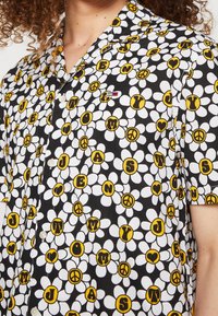 Camicia con maniche corte e bottoni, caratterizzata da uno sfondo nero con margherite bianche e accenti circolari gialli con lettere e simboli di pace.