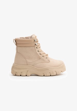 Botín de ante beige con cordones, cuello acolchado, suela gruesa y resistente, y tirador en el talón, mostrado en perfil lateral sobre fondo blanco.