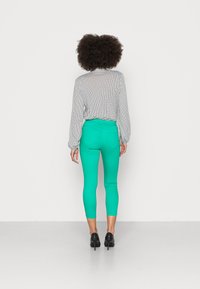 Leggings verde justos combinados com uma blusa de bolinhas pretas e brancas, com mangas longas e uma bainha arredondada. Sapatos de salto altos pretos completam o visual.