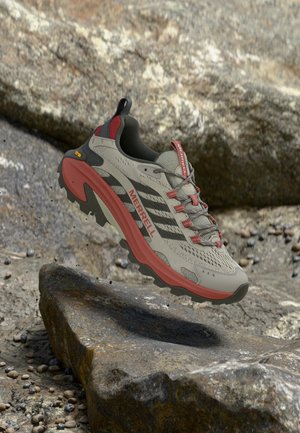 Scarpa da trekking Merrell grigio chiaro e rossa che fluttua sopra un terreno roccioso con suola robusta progettata per l'uso su sentieri all'aperto.