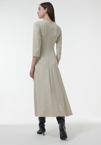 Un vestido largo de color beige claro con mangas tres cuartos, líneas limpias, un cierre oculto en la parte posterior y una silueta fluida. Llevado con botas negras hasta la rodilla.