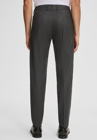 Uomo che indossa pantaloni da abito grigio a righe e scarpe nere, in piedi con le mani rilassate ai lati su uno sfondo semplice.