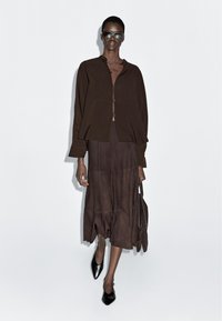 Brązowa kurtka zapinana na guziki z długimi rękawami, w zestawie z pasującą spódnicą do kolan. Modelka nosi ciemne okulary przeciwsłoneczne i czarne szpiczaste buty.