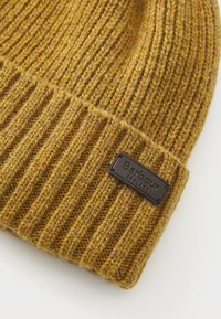 Căciulă din tricot cu rădăcini, de culoare muștar, cu margine pliată și etichetă neagră Barbour England cusută pe margine.