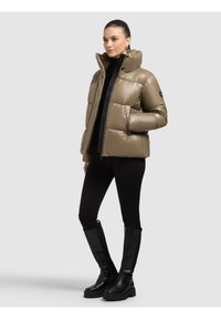 Kobieta w taupe pikowanej kurtce, czarnych legginsach i czarnych wysokich butach, stojąca bokiem na jednolitym jasnoszarym tle.