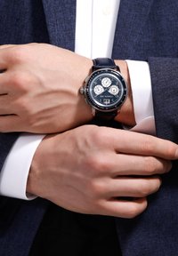 Reloj de hombre con correa de cuero azul oscuro, caja plateada, esfera azul, tres subesferas y un indicador de fecha; llevado en una muñeca con manos visibles.