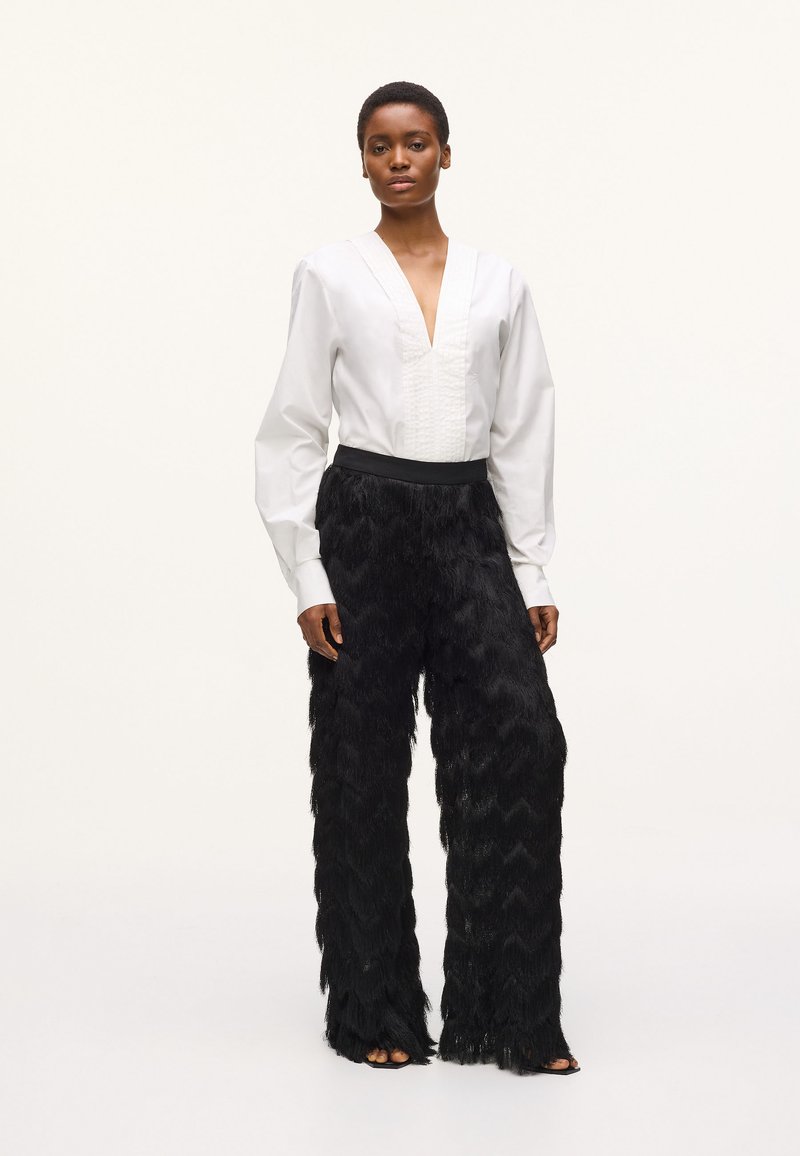 Blouse blanche à manches longues avec col en V et devant texturé, associ ée à un pantalon large noir à franges avec une taille lisse.