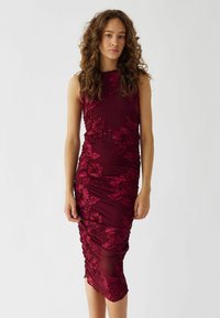Burgundyröd kroppsnära klänning med blommönstrad broderi och detaljer i rynk. Ärmlös design, knälång och slät textur med intrikata mönster.