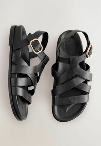 Sandales en cuir noir avec des brides croisées et une boucle latérale. Texture lisse avec une semelle plate et un design minimaliste.
