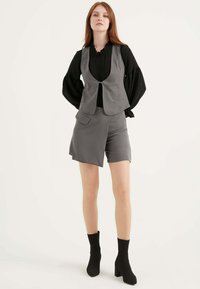Graue Weste und Shorts-Outfit aus glattem Material, mit Taschen, getragen mit einem schwarzen, Volant-Oberteil mit langen Ärmeln und schwarzen, hohen Stiefeln.