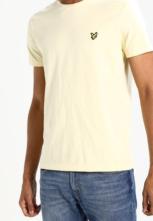 Mężczyzna ubrany w bladoróżową, krótkorękawową koszulkę z małym wyszywanym żółto-czarnym logo ptaka na piersi, w połączeniu z niebieskimi dżinsami.