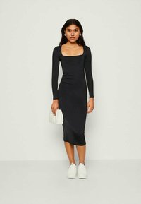 Abito bodycon nero a maniche lunghe con scollatura quadrata e dettagli arricciati, abbinato a una borsa bianca e a sneakers bianche. Tessuto morbido.