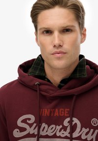 Sudadera con capucha granate con el logo "SuperDry", cordones y una camisa a cuadros verde debajo. Textura suave y diseño casual.