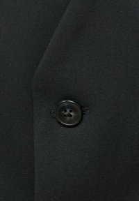 Crni materijal s glatkom teksturom sadrži zaobljeni, tamni gumb s četiri rupice za šivanje. Krupni plan detalja odjeće.