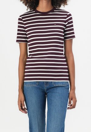 Persoon die een donkerpaarse en witte horizontaal gestreepte T-shirt en blauwe spijkerbroek draagt, staand tegen een effen witte achtergrond.