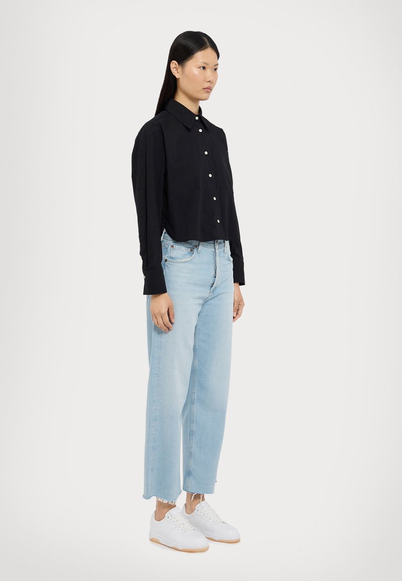 Camicetta nera corta con bottoni e maniche lunghe, abbinata a jeans azzurri a gamba larga con orli sfrangiati e sneakers bianche.
