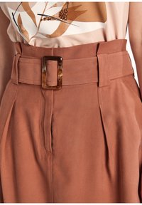 Pantaloni rustici a vita alta di colore marrone ruggine con cintura in tessuto e fibbia rettangolare in tartaruga, indossati con una maglietta beige a fantasia.