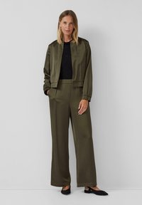 Vrouw die tegen een grijze muur staat, gekleed in olijfgroene wijdvallende broek, bijpassende jas, zwart top en zwarte slingback flats.