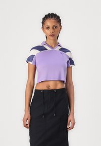 Polo crop em roxo com colarinho, apresentando padrões curvos em azul escuro e creme. S saia cargo preta com bolsos laterais e cordão.