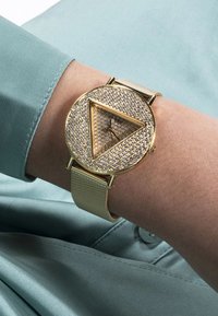 Guldfarvet ur med ansigt i triangulær form, dækket af små rhinsten, og en blød mesh-rem. Baggrunden har et lyseblåt stof.