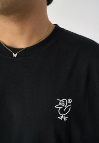 Schwarzes Baumwoll-T-Shirt mit einem weißen Cartoon-Vogel-Logo auf der linken Brustseite, kombiniert mit einer goldenen Schmetterlingsanhänger-Halskette.
