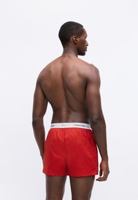 Rdeče kopalne hlače z elastičnim belim pasom, na katerem je napis "Calvin Klein", lahka tkanina in sproščen kroj.