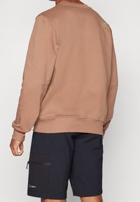 Sudadera marrón con puños y dobladillo acanalados, que presenta un pequeño logo en la parte posterior. Combinada con pantalones cortos cargo azul marino, uno de ellos con un bolsillo con cremallera.