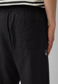 Pantalon en lin noir avec une poche arrière et une taille élastique. Une petite étiquette de marque est visible près de la couture de la poche.
