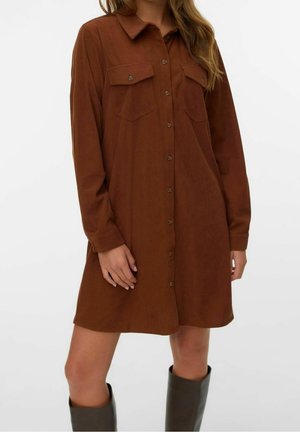Femme portant une robe-chemise marron à manches longues avec poches poitrine à rabat, associée à des bottes hautes foncées.