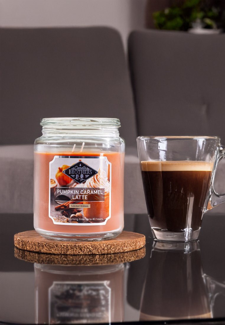 Bougie en bocal en verre étiquetée "Pumpkin Caramel Latte", cire de couleur pêche, couvercle transparent, sur un sous-verre en liège avec un verre de café noir.