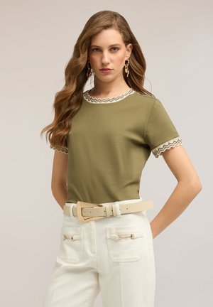 Donna che indossa una maglia verde oliva con maniche e collo bordati a zigzag, pantaloni bianchi a vita alta con tasche frontali e una cintura beige.