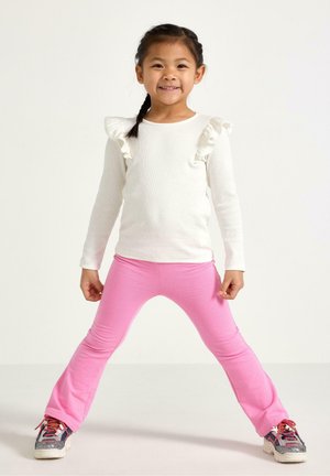 Ung pige står med benene spredt, iført hvid bluse med volanter og lange ærmer, lyserøde bukser og sneakers, smilende til kameraet.