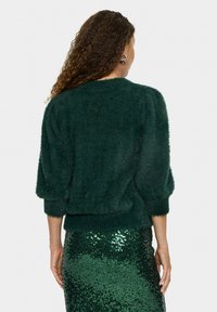 Dunkelgrüner, flauschiger Pullover mit puffigen Ärmeln und geripptem Saum. Getragen mit einem glitzernden grünen Rock, der die Textur und den Farbkontrast hervorhebt.