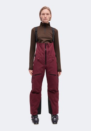 Un mannequin se tient devant un fond uni, vêtu d’un haut marron, d’un pantalon de ski à bretelles bordeaux et de chaussures de ski noires.