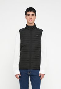 Čierna prešívaná vesta s vysokým golierom, predným zipsom a bočnými vreckami. Má logo na hrudi a textúrované panely na pleciach.