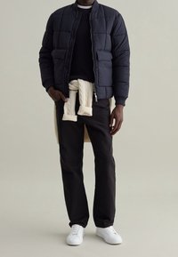Giacca bomber trapuntata blu navy con due tasche frontali, indossata sopra un maglione scuro. Pantaloni neri e sneakers bianche completano il look.