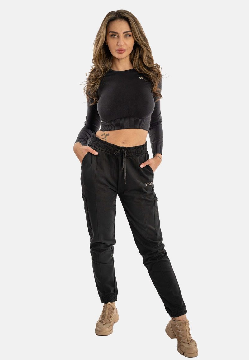 Schwarzes, kurzes Sweatshirt mit langen Ärmeln und schwarzen Jogginghosen, die Taschen und elastische Bündchen haben. Hellbeige Sneakers vervollständigen das Outfit.