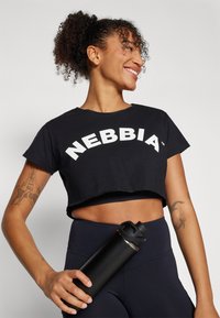 Crni kratki majica s bijelim tekstom "NEBBIA", u kombinaciji s crnim hlačama visokog struka. Model drži elegantnu crnu bočicu za vodu.