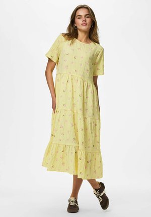 Femme portant une robe midi jaune ample à manches courtes, broderies florales et baskets marron, debout sur un fond blanc uni.