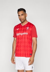 Červený futbalový dres s diamantovým vzorom, bielymi akcentmi a okrúhlym výstrihom. Na prednej strane a rukávoch sa nachádzajú logo a branding.
