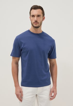 Man met kort, donker haar en baard die een effen blauw T-shirt met korte mouwen en witte broek draagt, staand tegen een effen achtergrond.