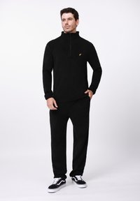 Juodas vilnonis džemperis su pusiau užtraukiamu kaklu ir auksiniu logotipu, derinamas su atitinkančiomis juodomis kelnėmis ir juodais sportbačiais su baltais akcentais.