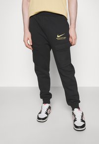 Svarta cargo-joggingbyxor med sidofickor, med en gul Nike-logotyp tryckt på höften. Tillverkade av bomull, har de en avslappnad passform och avsmalnande ben.